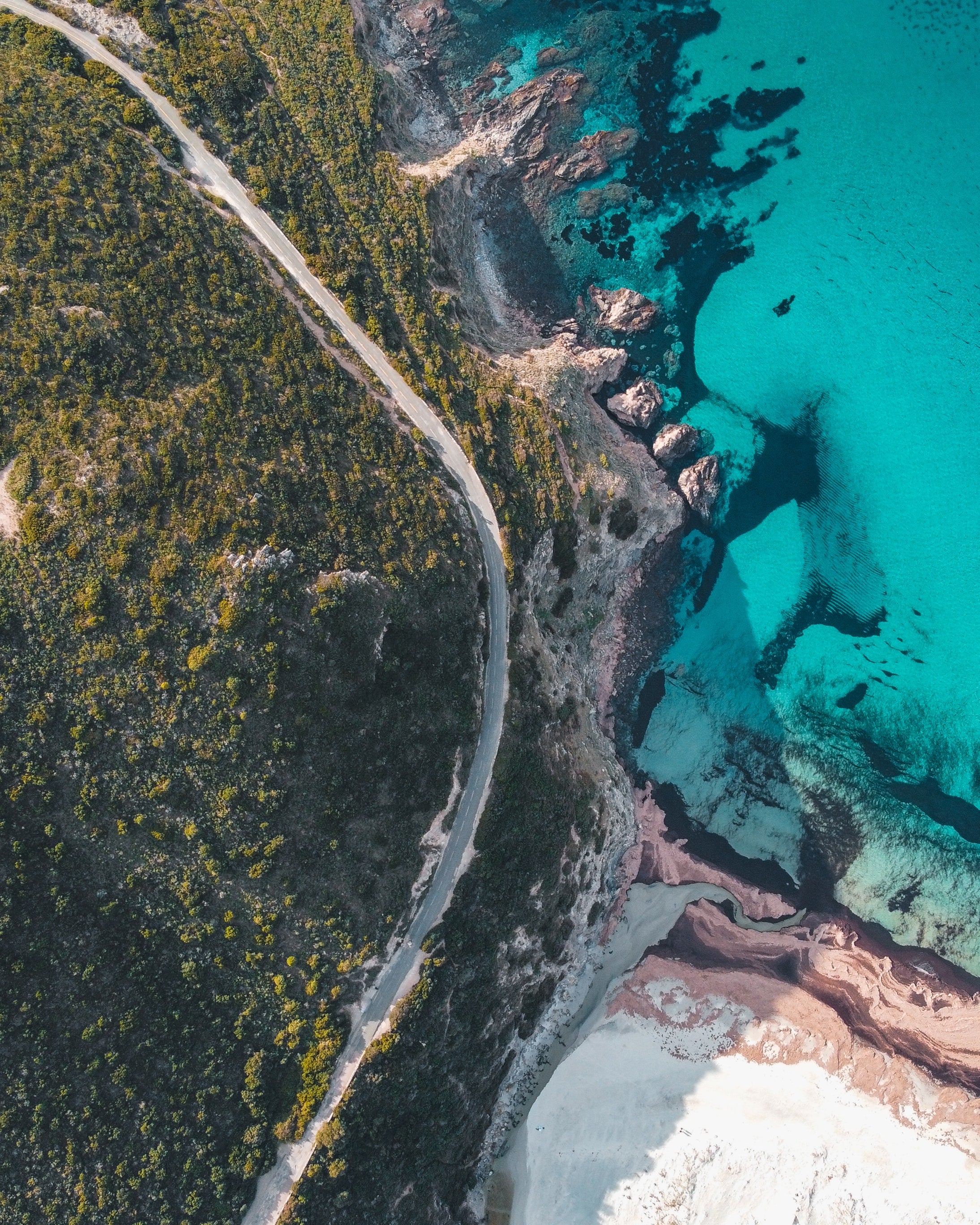 Magnifique photo aérienne de la plage de l'Ostriconi en Corse. On y voit la mer turquoise, la plage de sable blanc, une route côtière sinueuse et le maquis verdoyant. 