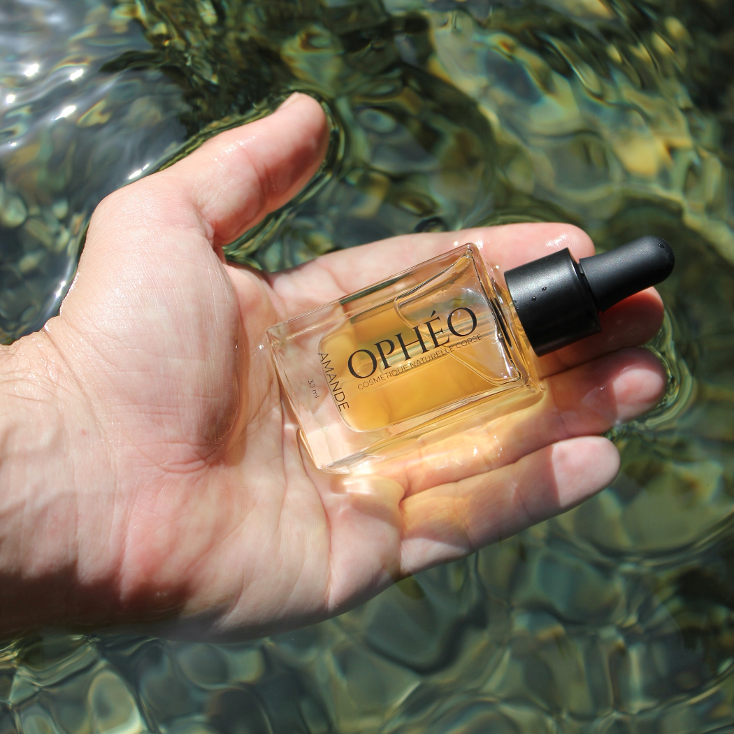 Un flacon d'Huile Visage Amande Nourrissante de la marque OPHÉO est tenu par une main d'homme au dessus de l'eau calme d'une rivière corse. L'eau est d'un magnifique vert émeraude et le soleil vient se refléter sur l'eau.