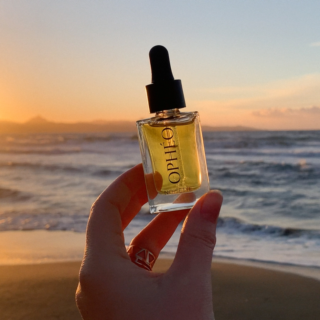 Un flacon d'Huile Visage Noisette Régulatrice de la marque OPHÉO est tenu par une main de femme. En arrière-plan, une plage et la mer corse au coucher du soleil. Le ciel est orange et bleu.