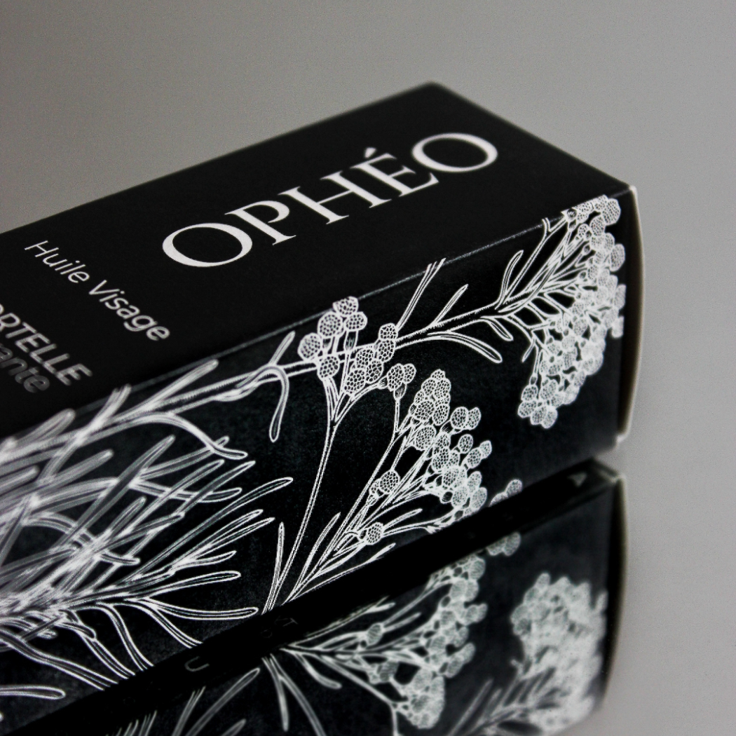 L'emballage carton de l'Huile Visage Immortelle Régénérante de la marque OPHÉO. Il est noir et blanc, d'un style élégant et épuré. On y voit une magnifique illustration botanique représentant une plante : l'immortelle corse.