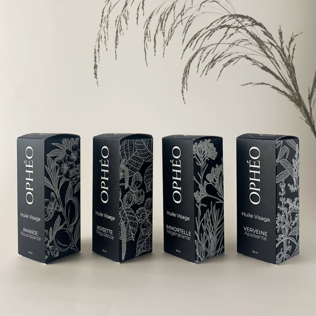 Les quatre Huiles Visage OPHÉO sont alignées dans leur emballages carton noir et blanc. Les emballages carton sont d'un style élégant et raffiné. Sur le côté de chaque emballage carton, on peut apercevoir une illustration botanique de la plante composant l'ingrédient principal du soin.