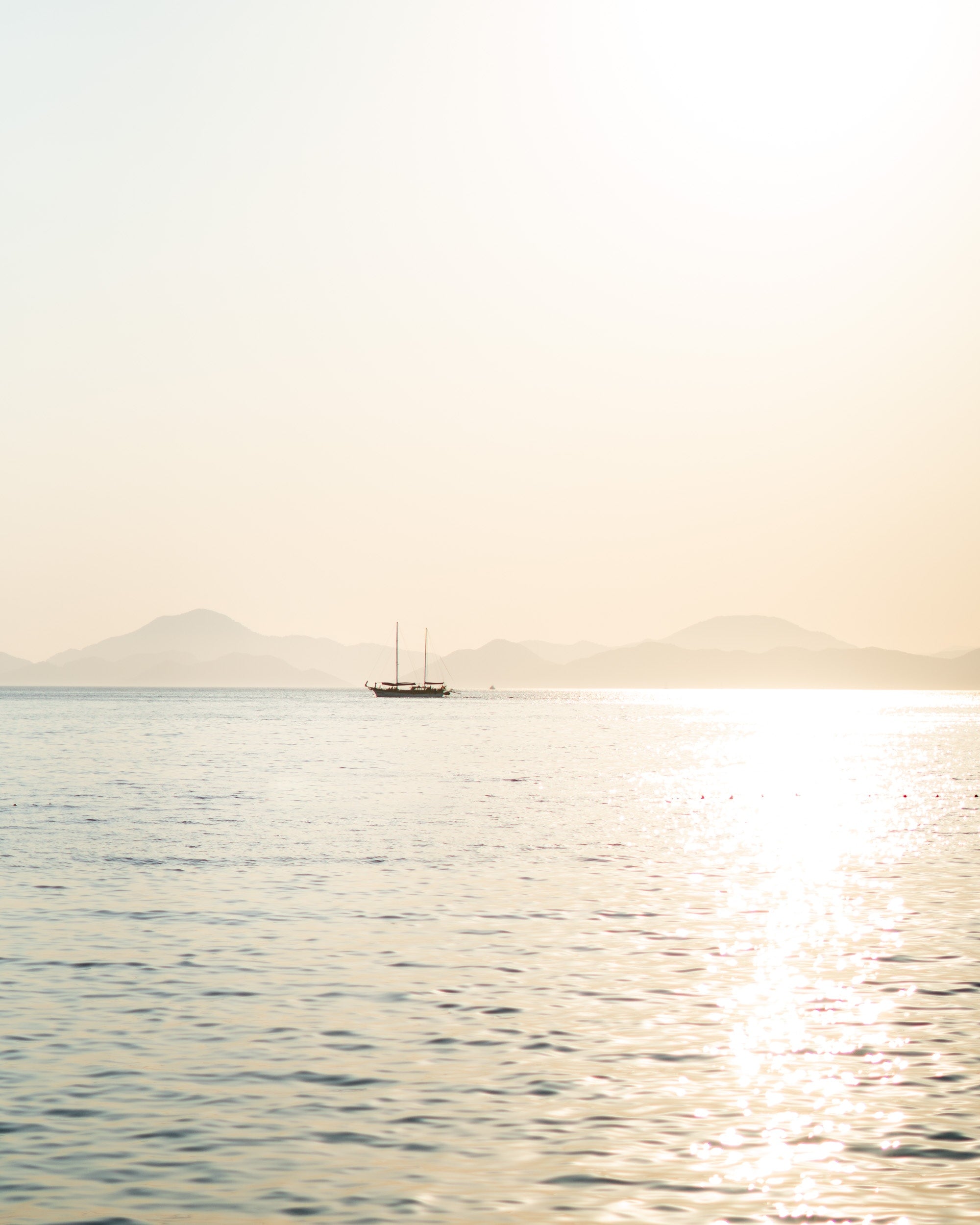 Photo d'une mer bleu ciel et scintillante sous un soleil doux et doré. Un voilier et des montagnes sont visibles à l'horizon.
