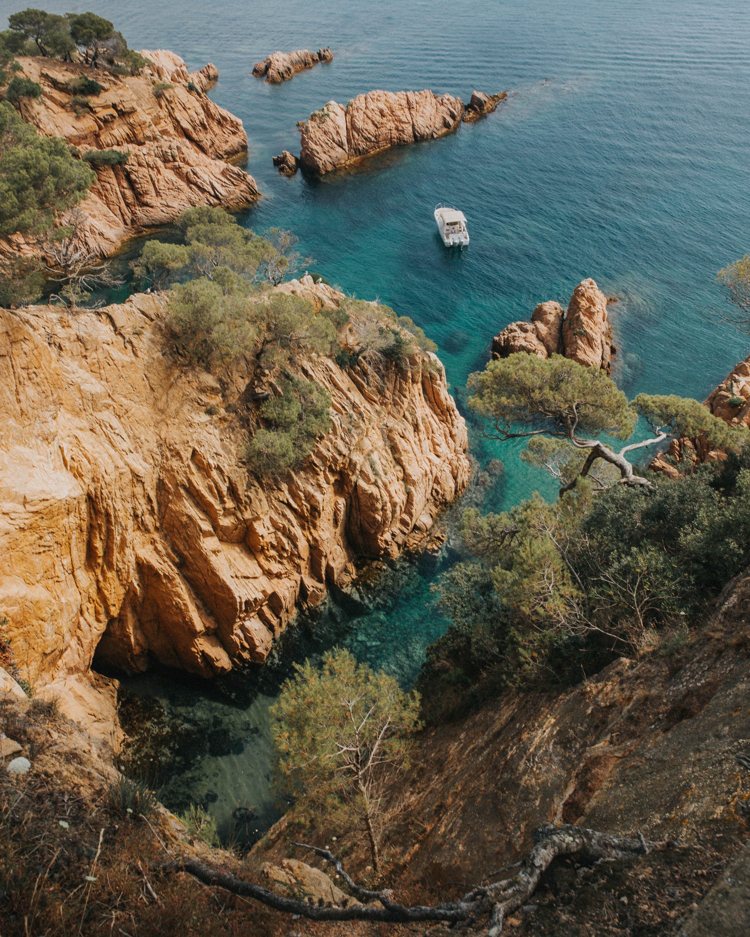 Photo montrant la côte méditerranéenne: falaises rouges, eau turquoise, criques avec un bateau, et pins.